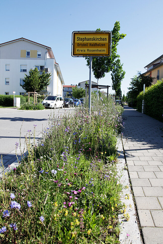 Eine Verkehrsinsel mit artenreicher, blühender Bepflanzung vor dem Ortsschild der Gemeinde Stephanskirchen ist zu sehen.